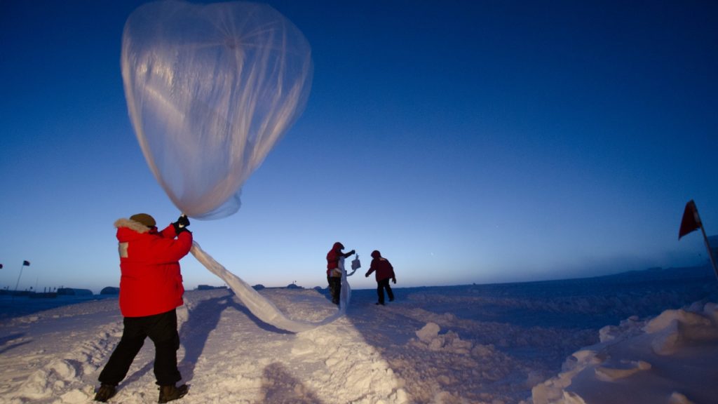 Person wearing thick orange coat holds a large balloon filled with helium, they stand on snowy ground.