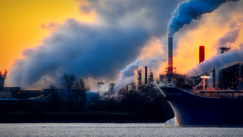 Large ship moves in front of factories on the shoreline, large smoke stacks rise from the ship and the factory