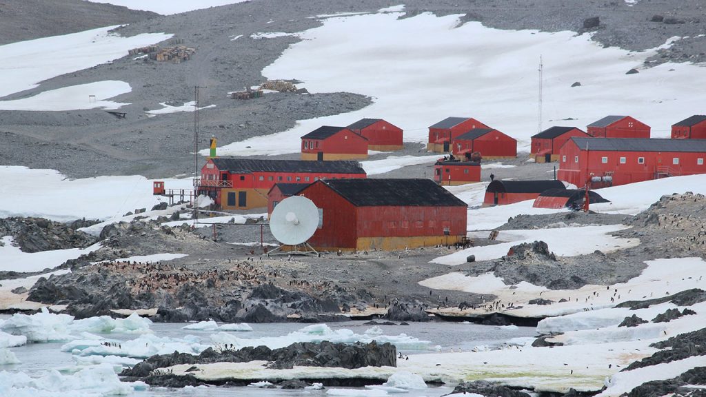 Red buildings on grey, rocky land, with patches of ice nearby. There is a white, circular dish in the centre.