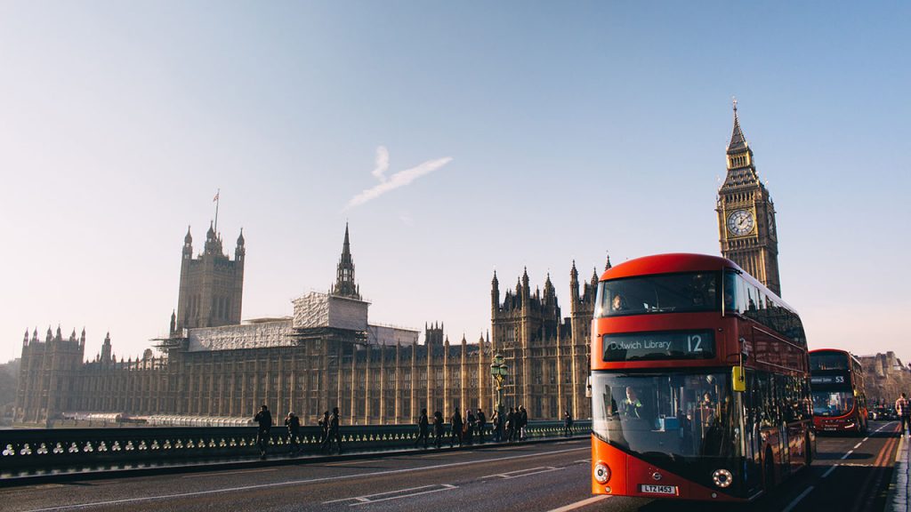 Red London bus moves along a quiet road. Large yellow brick buildings in the distance, including Big Ben.