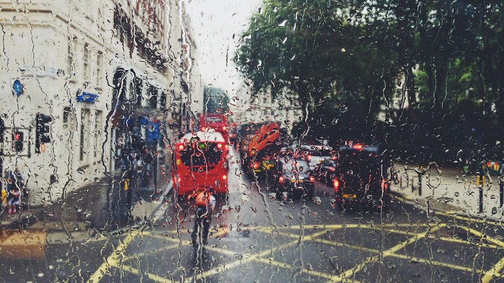 Red bus and man wearing orange jacket riding a bike travel down a road, rain droplets distort the view