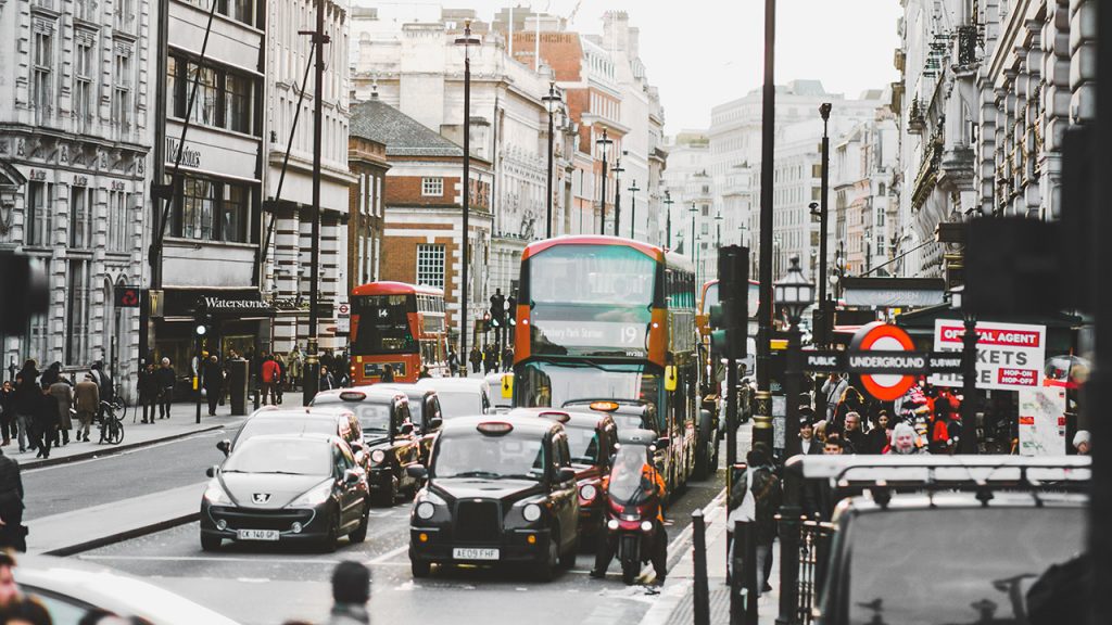 Cars and buses queue at traffic lights in London, next to underground tube station sign.