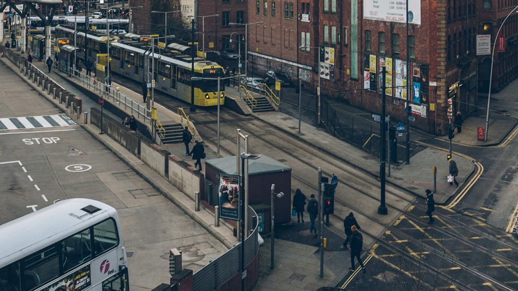 Tram moving from left to right through a busy city centre, with many people walking along the pavement.