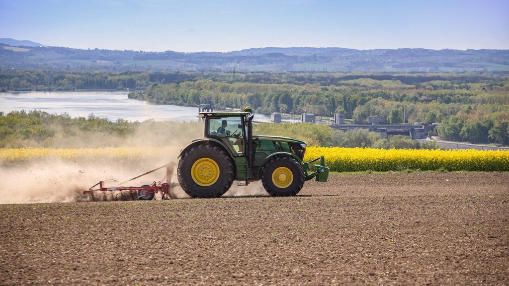 Green tractor ploughs a dusty brown field, with green trees behind.