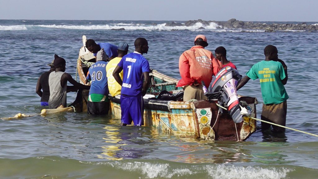 Nine men stand in shallow water offshore, surrounding a small fishing boat with a motor at the rear.