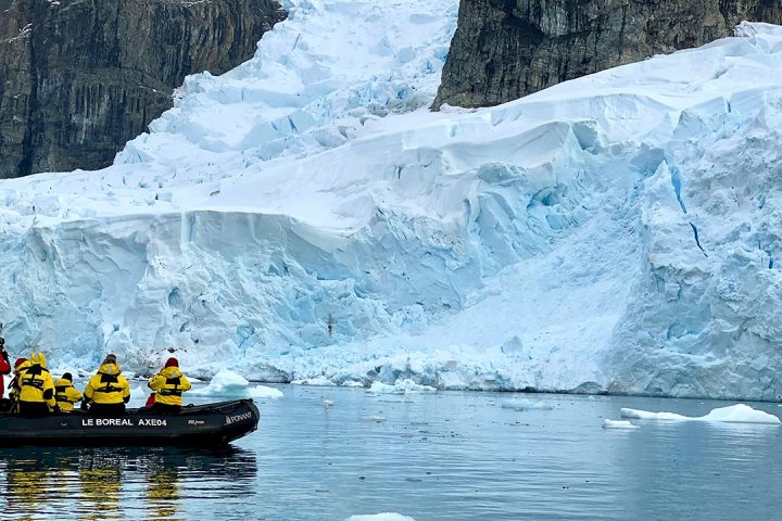 Team of people wearing yellow jackets stand on a small black powerboat, they are looking at white sheets of ice in the ocean.
