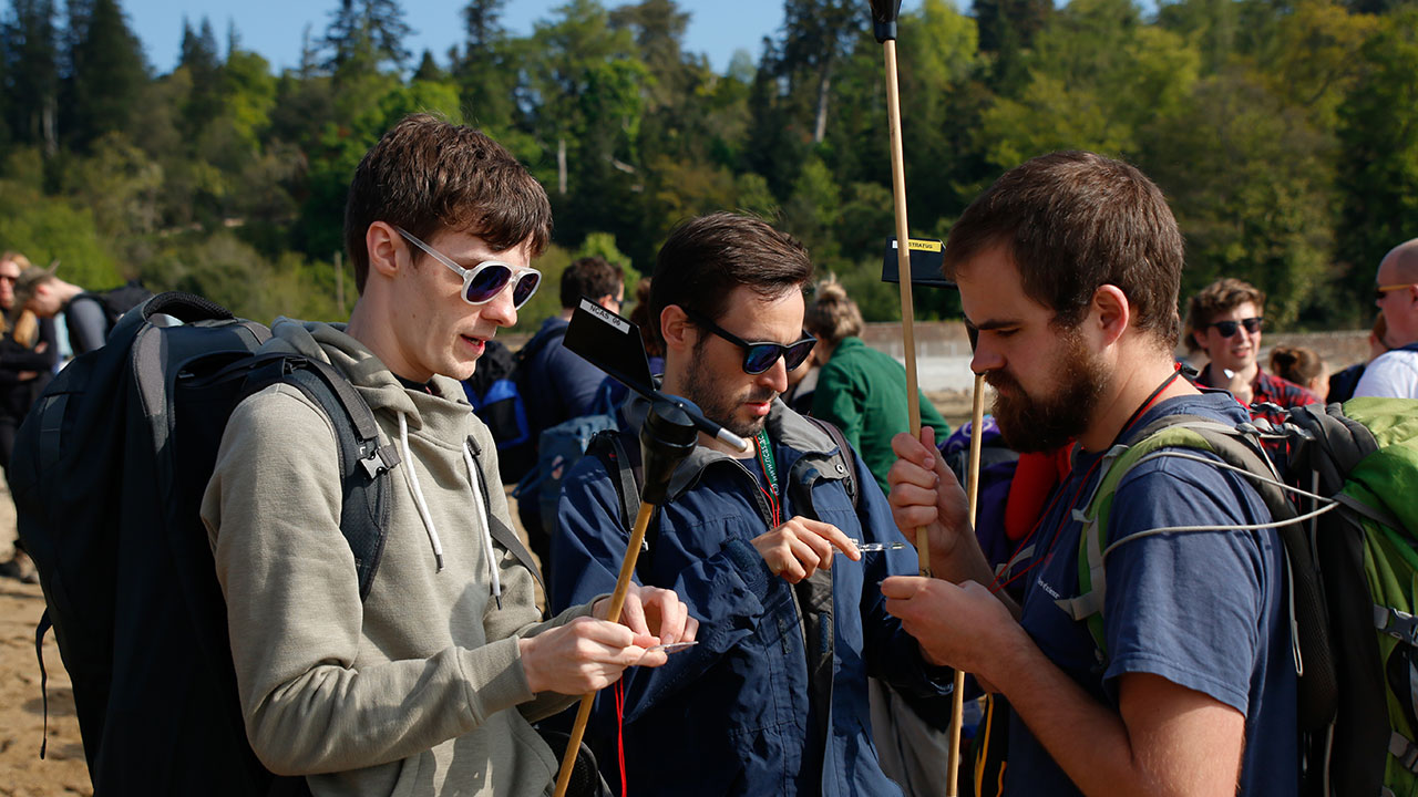 Three men, two wearing sunglasses, look closely at small scientific instruments in their hands