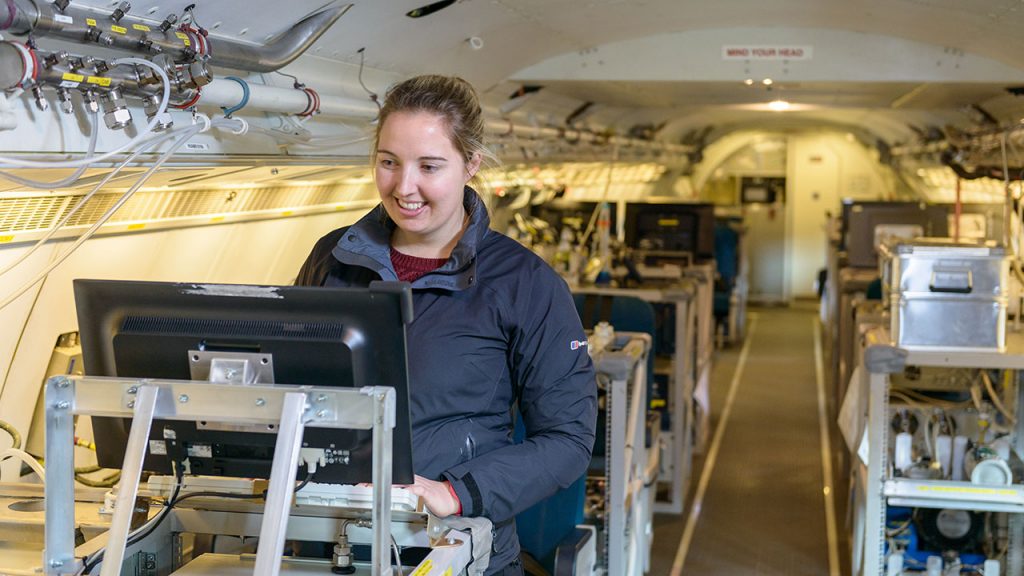 Woman wearing blue jacket stands at a computer desk inside an aircraft full of scientific kit