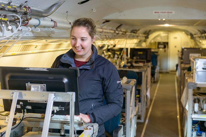 Woman wearing blue jacket stands at a computer desk inside an aircraft full of scientific kit