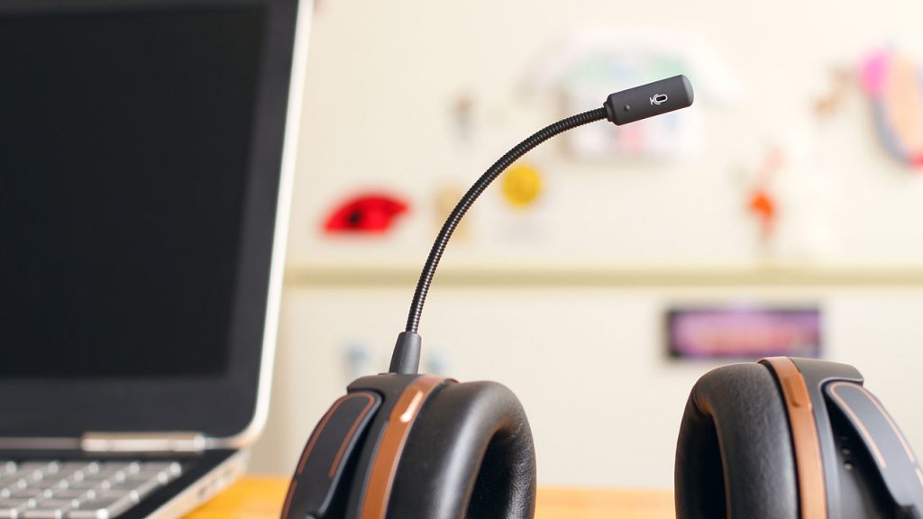 Black headphones with mouthpiece rest on a wooden desk, next to a laptop.