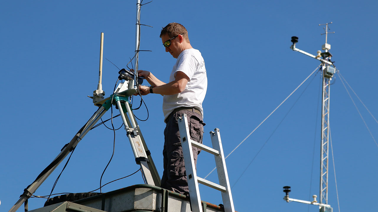 Man wearing grey t-shirt stands next to a ladder, he is using his hands to adjust a scientific instrument sitting on a metal tripod
