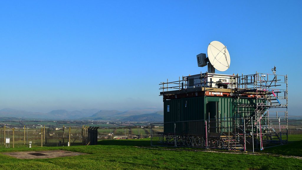 White radar dish sits on top green shipping container in a field, with blue skies behind