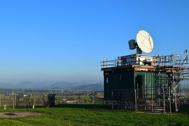 White radar dish sits on top green shipping container in a field, with blue skies behind