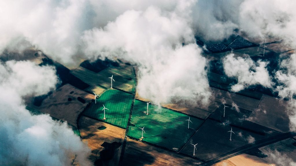 Green patchwork fields and wind turbines are seen through a layer of fluffy white clouds