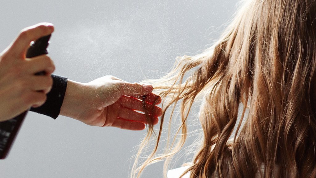 Person spraying hair product onto long, blonde hair