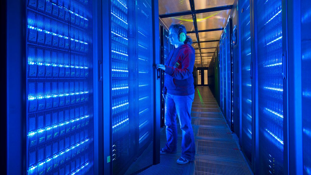 Woman wearing earphones stands in corridor between electric blue illuminated computer hardware