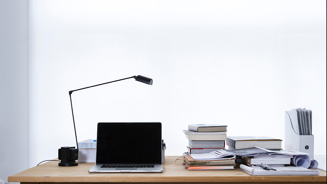 Wooden desk with laptop, reading lamp and piles of books.