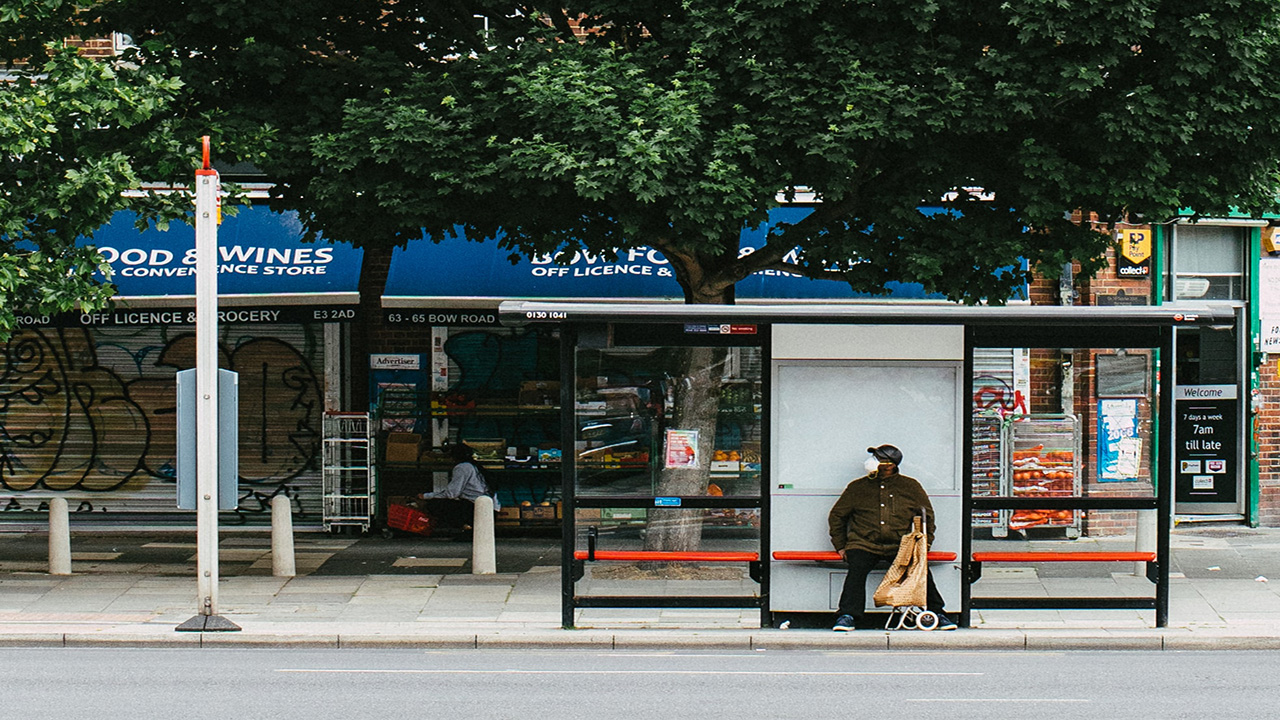 Man wearing a face mask, sat at a bus stop on a tree-lined street with shops