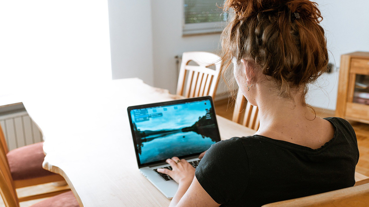 A woman with tied up curly hair, working on a laptop on a dining room table