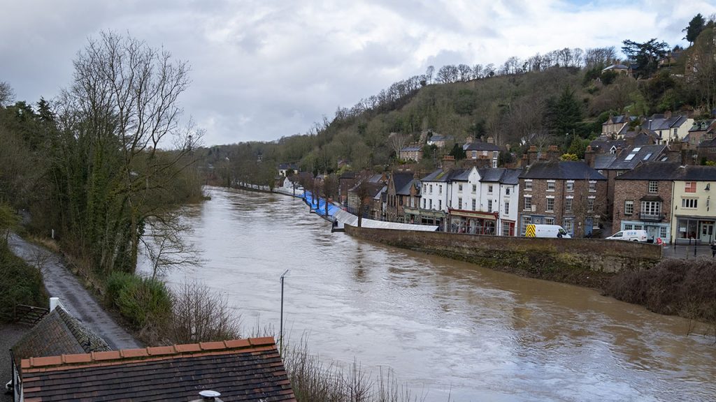 Brown river water flows through small village, with flood barriers along the road