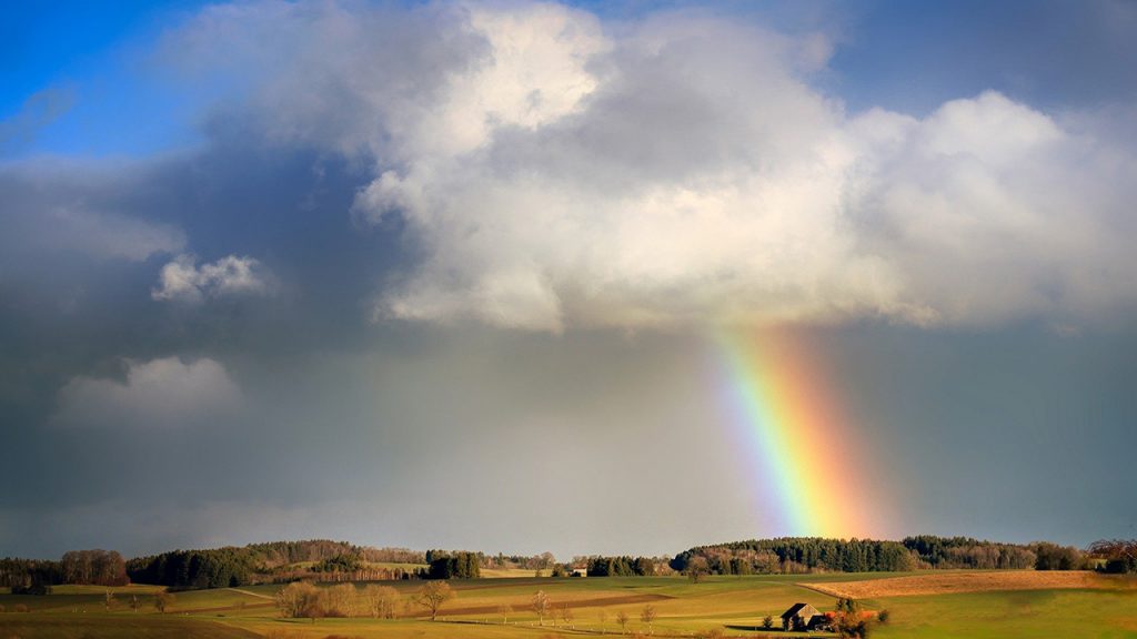 Cloudy blue sky with a rainbow, over a couple of fields and woodland