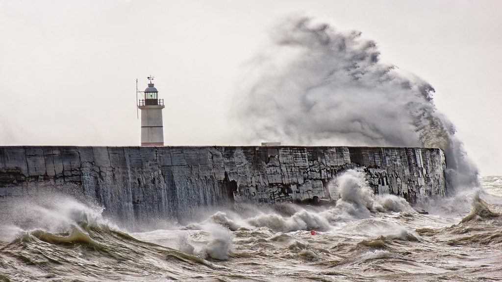 A wave hits a grey sea wall and sprays up in the air next to a lighthouse