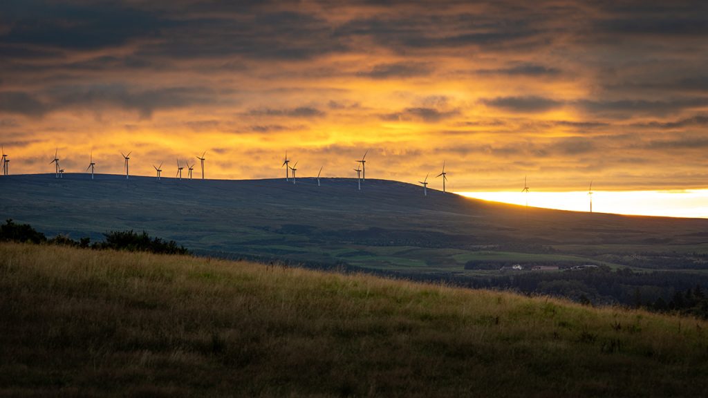 Rolling hills with wind turbines on the horizon, a sunset creates a vivid yellow sky.
