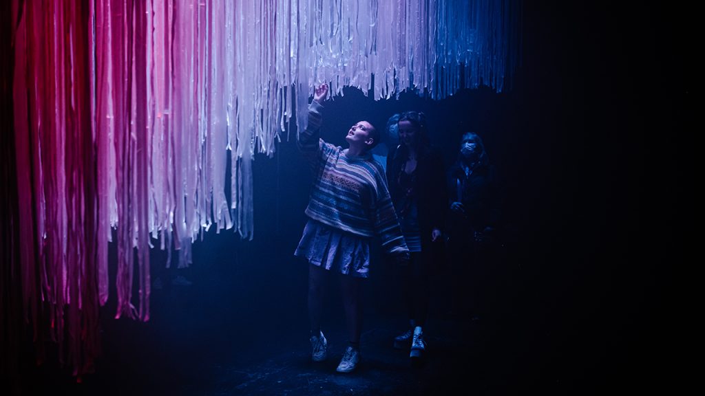 Three people reach up towards a hanging canopy of material, illuminated in blue and red