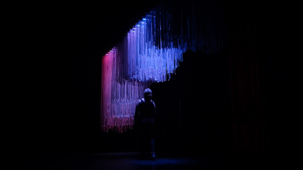 Person walks underneath hanging art exhibition. Twenty-six coloured stripes illuminate dangling plastic ribbons.