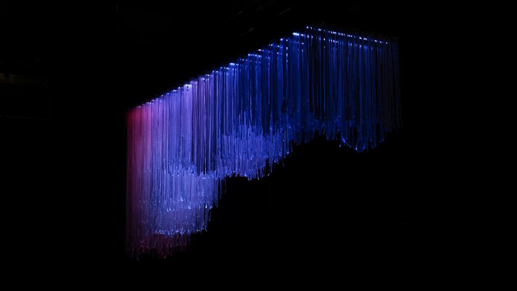 Twenty-six coloured stripes, illuminating hanging ribbons, suspended from above.