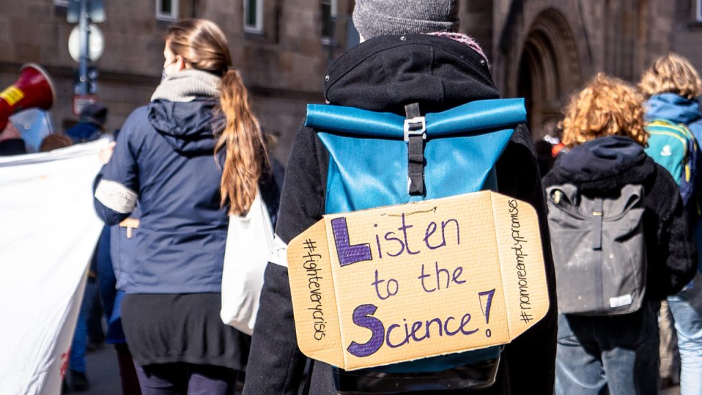 Person wearing blue backpack with sign reading "listen to the science"