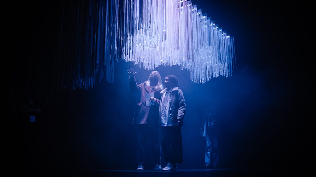 Two people stand underneath a hanging fabric canopy, illuminated in blue and red