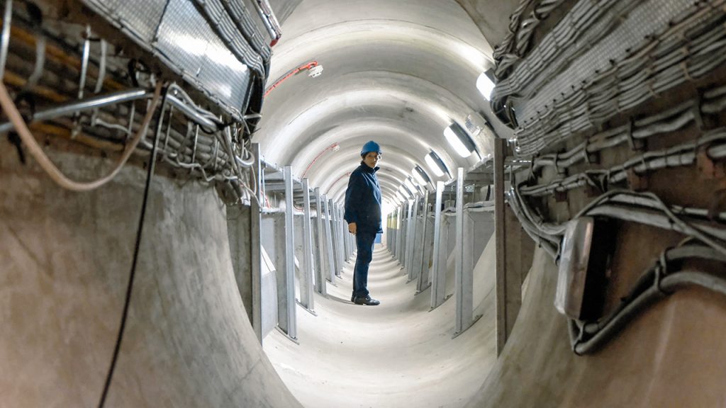 A woman wearing a hardhat, jeans and a dark jumper stands inside a grey tunnel with cables in the foreground