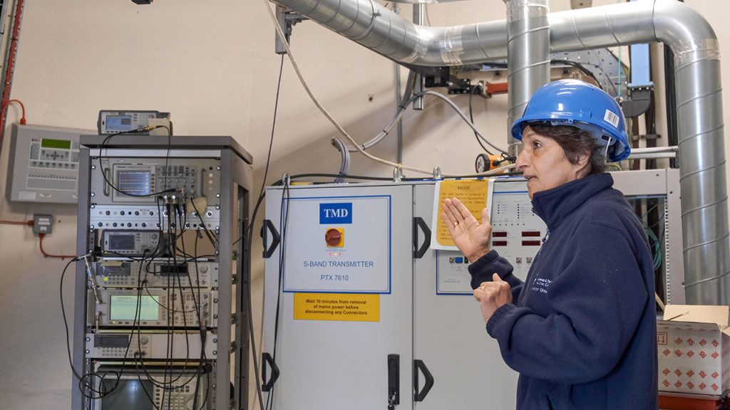 Scientist wearing a blue hard hat stands beside a radar signal transmitter