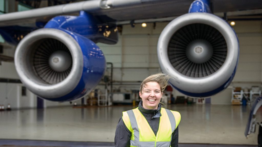 A woman in a hi-vis jacket stands smiling outside of an aircraft hangar