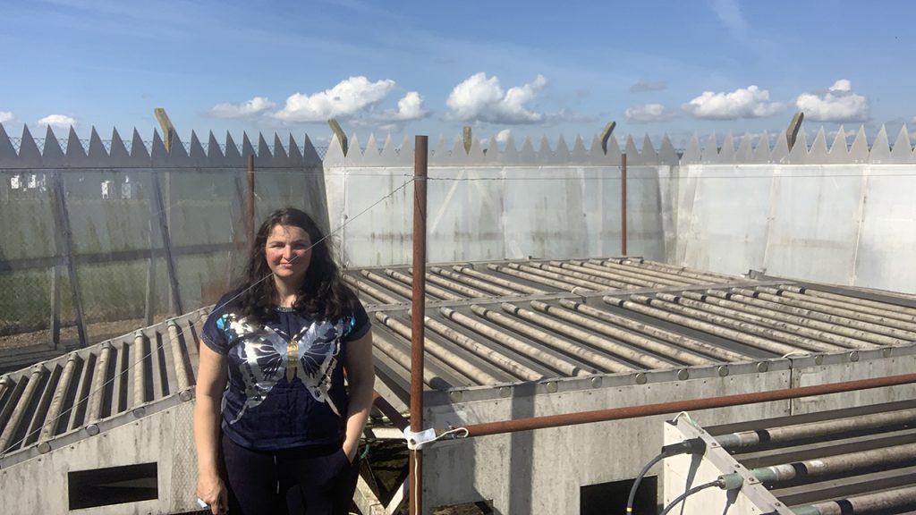 A woman with long dark hair stands on a roof next to a scientific instrument with a blue sky and grey rooftop fence