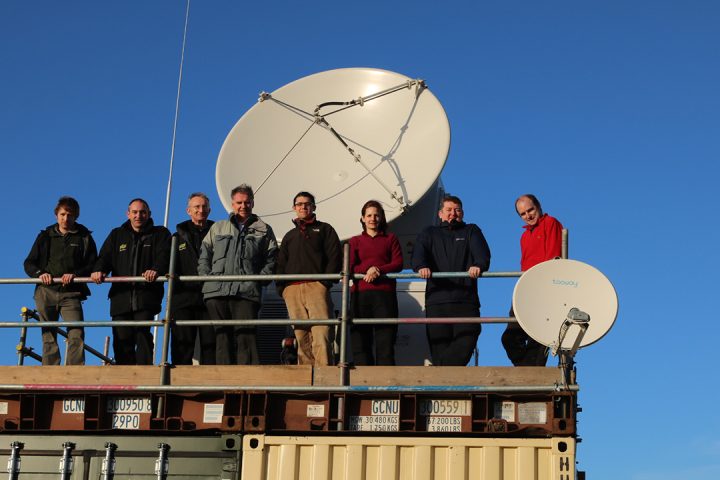 Eight people stand in front of a white radar dish, with a bright blue sky behind them