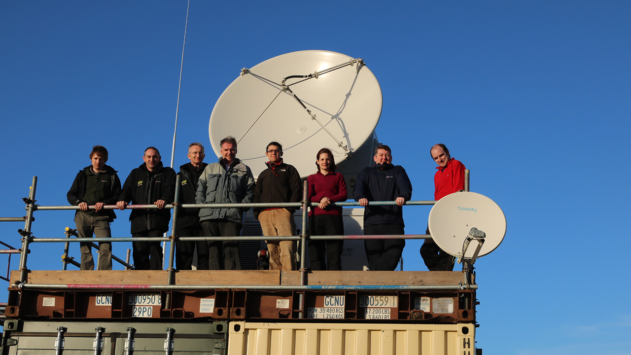 Eight people stand in front of a white radar dish, with a bright blue sky behind them