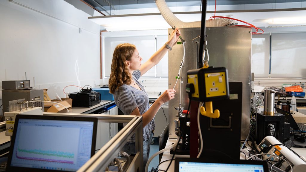 Person in laboratory space holds wrench and gas inlet tube on large metal chamber