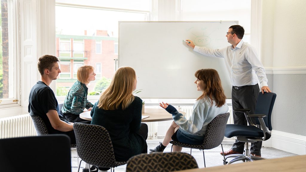 three women and one man sit at a round table looking at a standing man pointed to a whiteboard