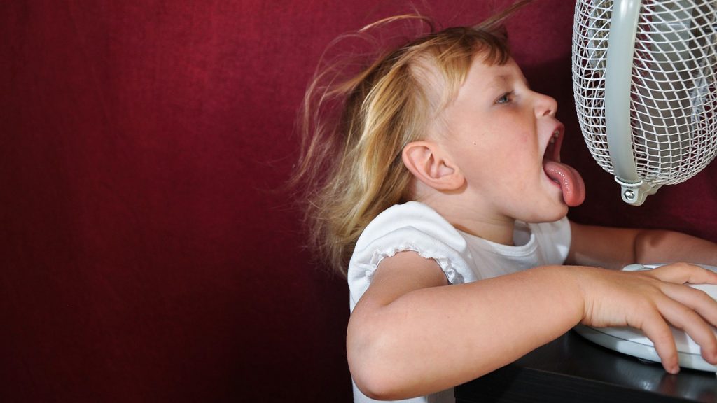 A small child with blond hair sticks out their tongue in front of a fan to cool down