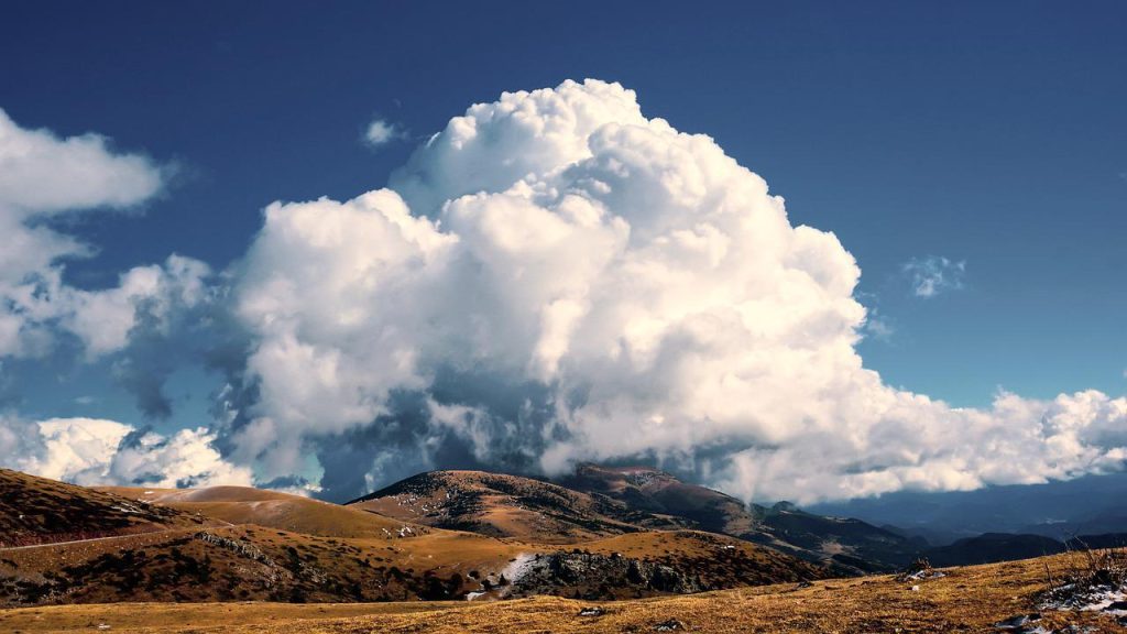 Large fluufy white clouds forming over the top of a brown mountainous landscape