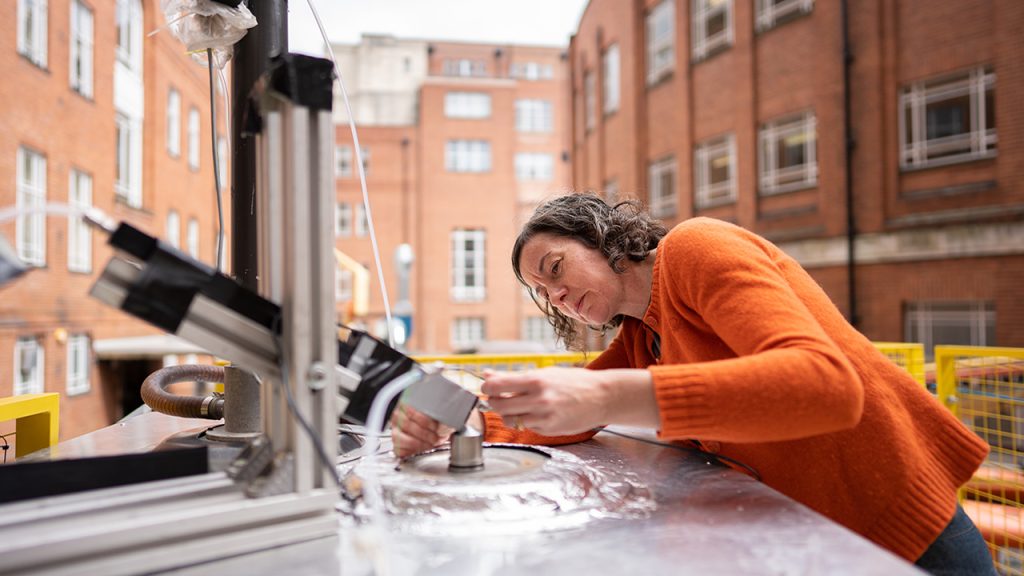 A woman wearing an orange cardigan looking at a piece of chemistry equipment outside, with brick buildings in the background