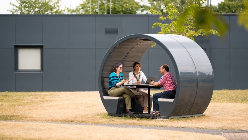 Three people sat inside a circular bench pod, outside by some dry brown short grass and green trees