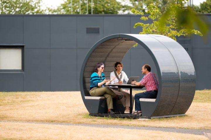 Three people sat inside a circular bench pod, outside by some dry brown short grass and green trees