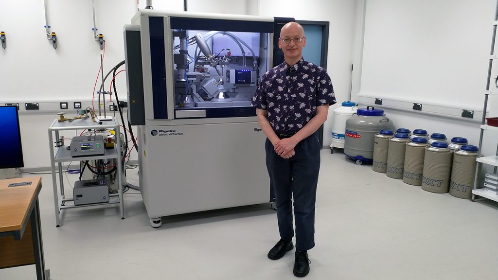 Professor Kevin Cowtan is stood in front of a crystallography piece of scientific equipment in a laboratory