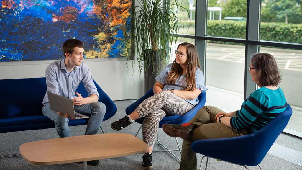 A group of people sat down having a conversation. One is holding a laptop.