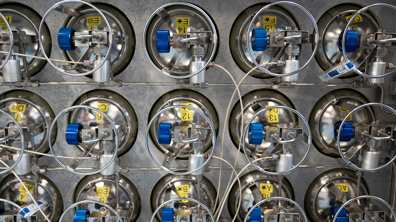 silver gas cylinders stacked in neat rows with blue dials, yellow labels, and white connected tubes