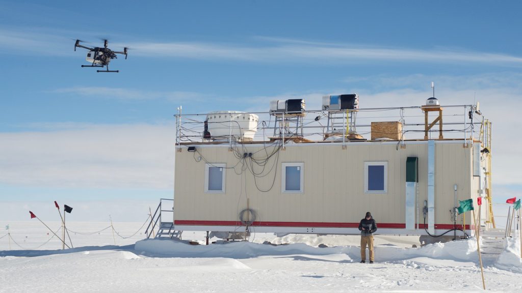 person stands on snow in front of a beige building while flying a drone