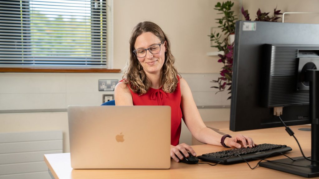 A woman sat at a desk looking at laptop with computer screen in front of her.